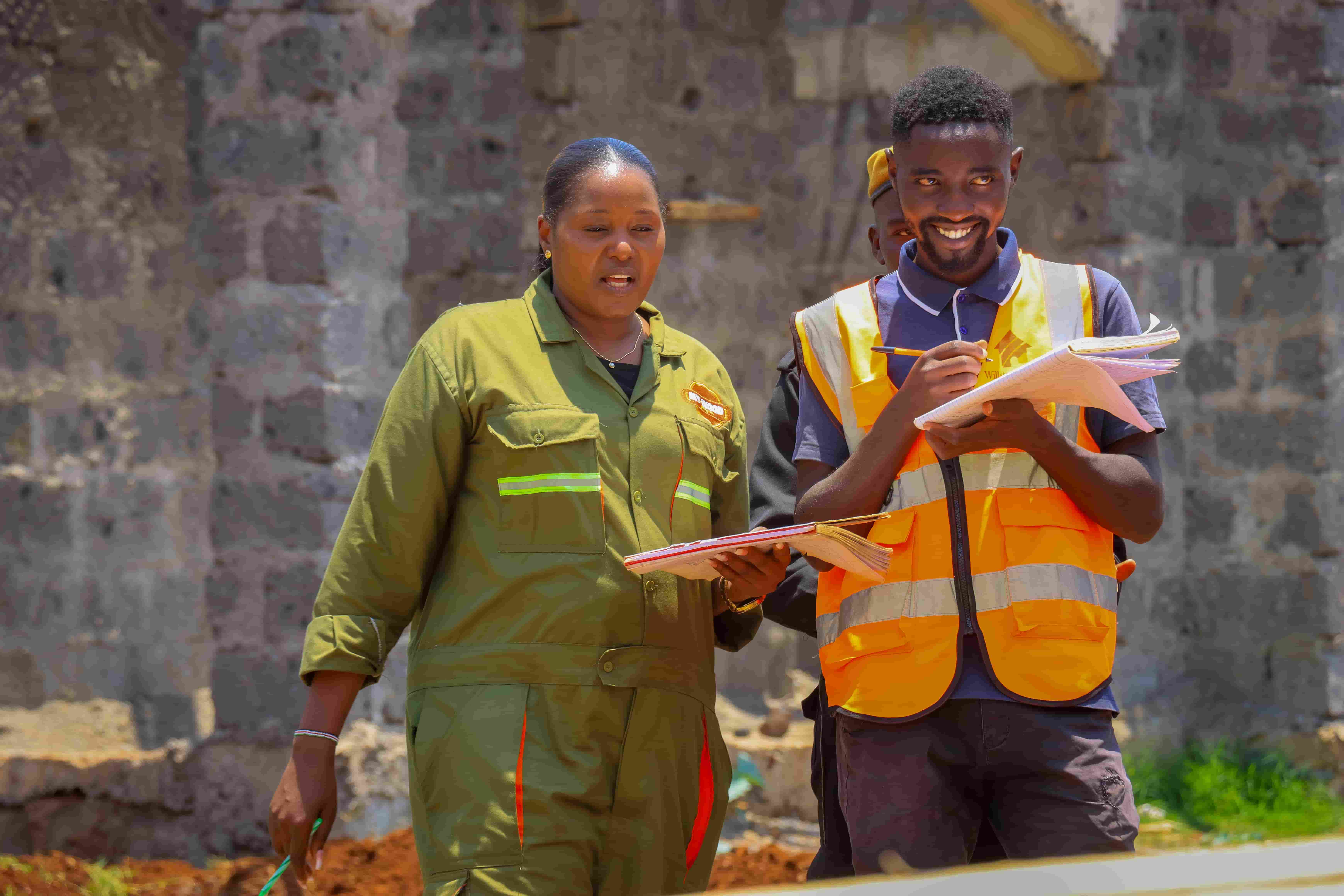 Two construction workers, a woman in a green coverall with reflective stripes and a man in an orange safety vest, are standing outdoors on a construction site. They are holding notebooks and appear to be reviewing or discussing plans. The background shows a partially constructed stone wall.