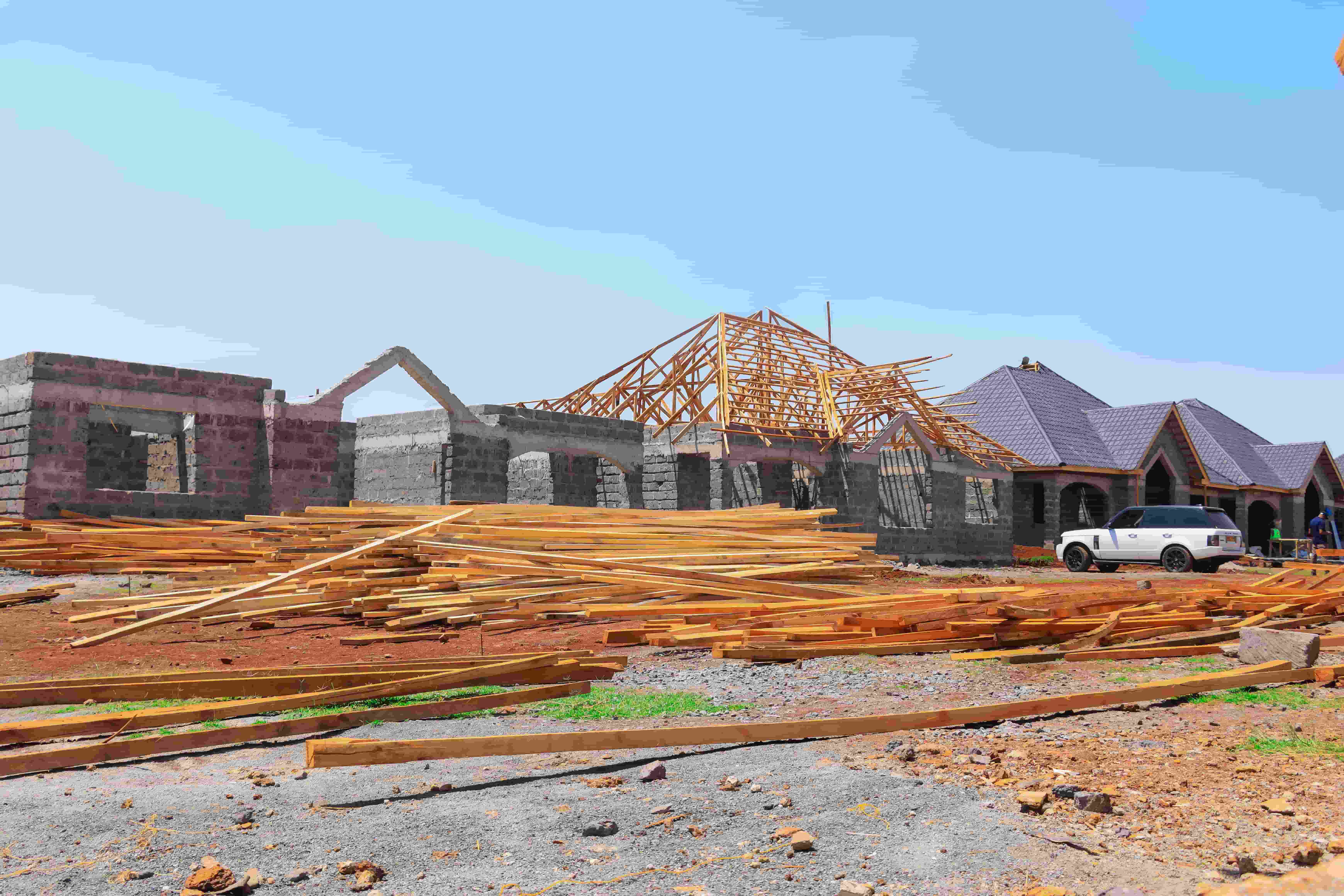 Residential house under construction with stone walls, wooden roof trusses, and timber planks on the ground next to a completed house.
