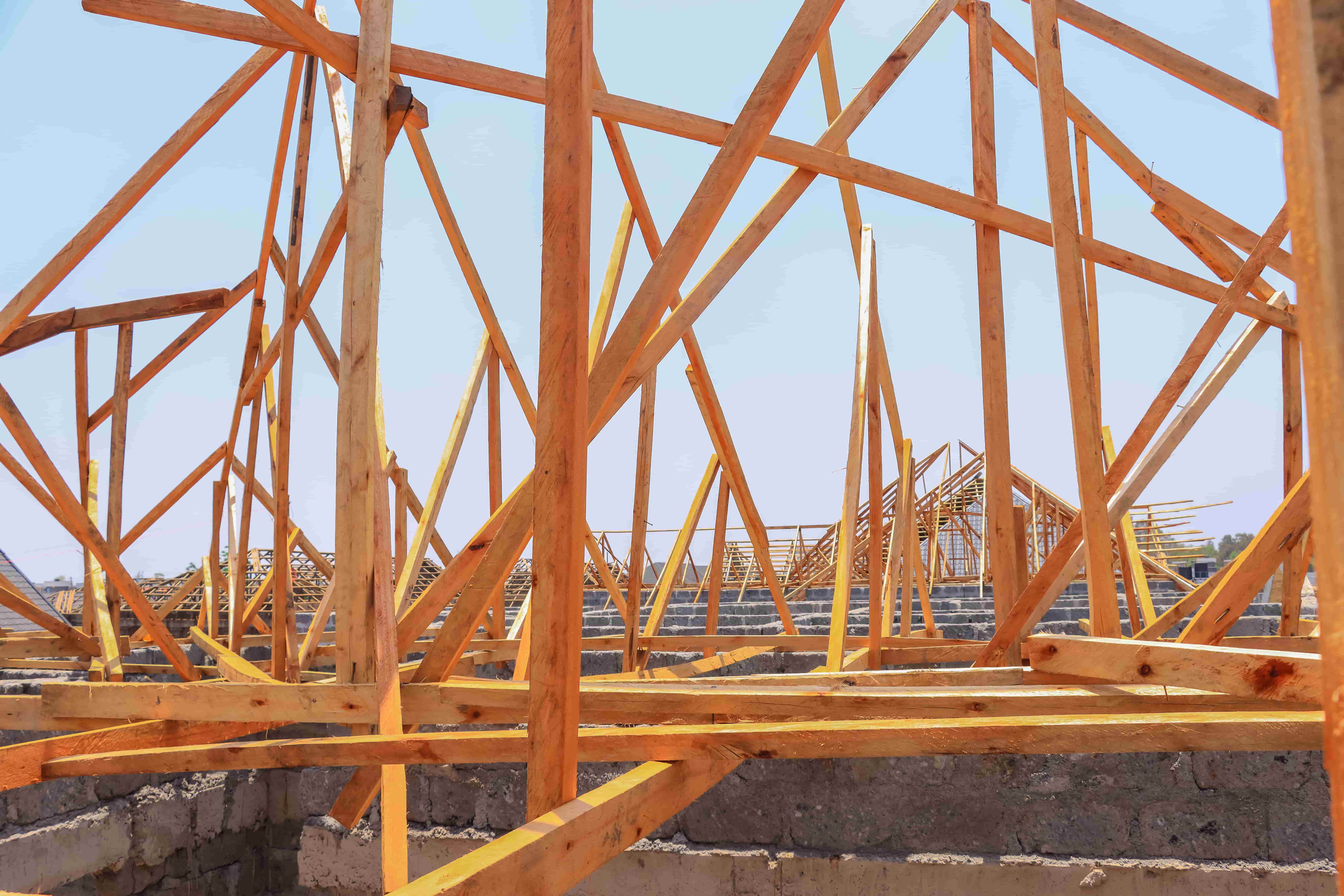 Timber roof trusses being installed on a building under construction in Kenya, showing wooden framework and stone walls under a clear sky.